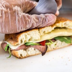 Hand slicing a delicious gourmet sandwich with meats, cheese, and greens.